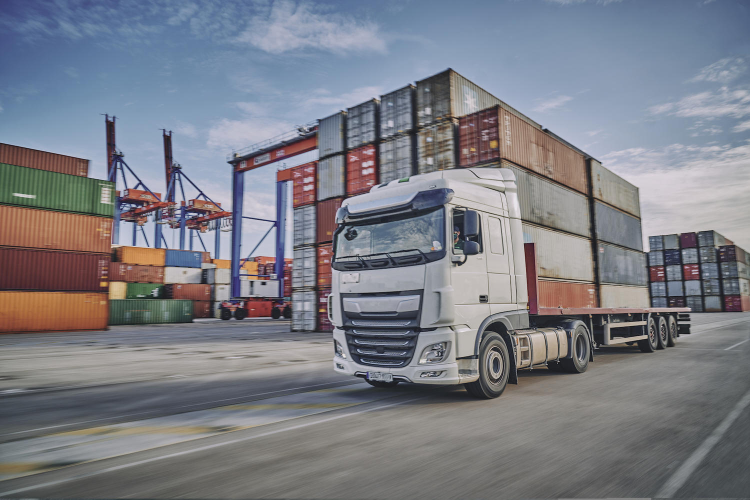 A white transport truck driving through a container terminal with stacked containers and cranes behind