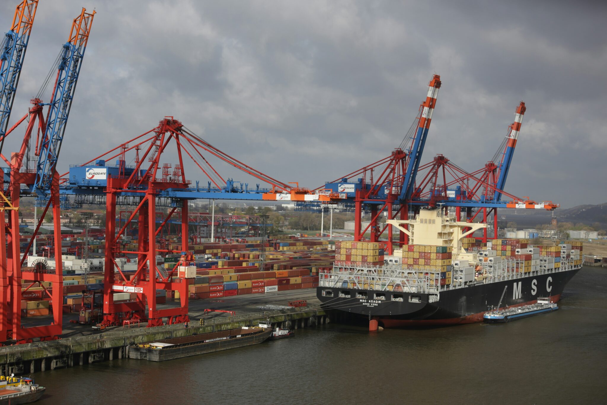A container vessel berthed under red and blue gantry cranes at a container terminal
