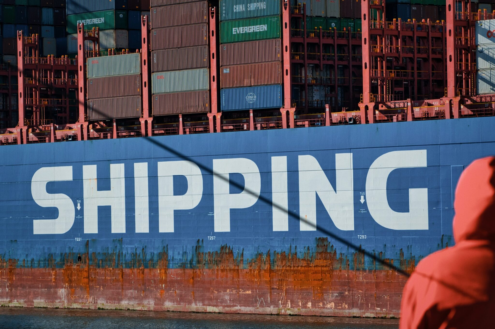 A container ship's hull lettered SHIPPING, stacked with containers, a dockworker in a red jacket in the foreground
